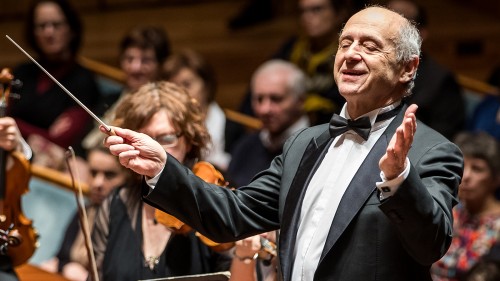 An older white man in a suit and a bow tie conducts an orchestra with arms outstretched in front of a crowd. 