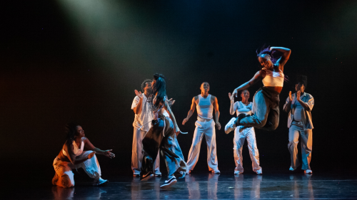 In blue light, three dancers in streetwear pose with legs apart, one foot lifted, and both arms gesturing to the side. 