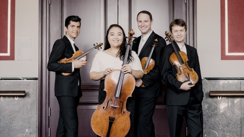 A string quartet of 3 women and a man stand in front of closed light door. 