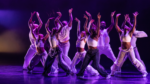 dancers in streetwear under purple stage lighting. They stand in strong poses with their legs apart and their arms raised
