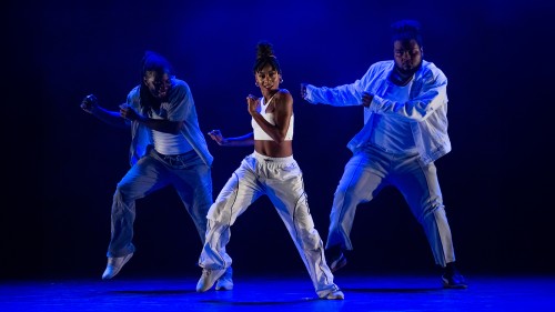 In blue light, three dancers in streetwear pose with legs apart, one foot lifted, and both arms gesturing to the side. 