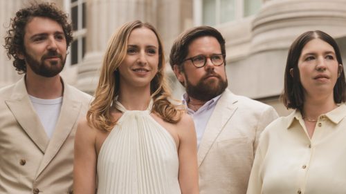 Four musicians stand smiling in front of a building with pillars in white and cream outfits. 