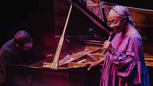 A black woman wearing a patterned durag, round glasses, and a purple flowing dress stands by a grand piano holding a microphone. 