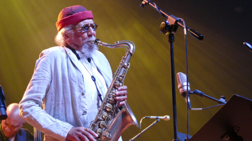 An older Black man, with a white goatee and wearing a knit cap and aviator sunglasses,  plays a tenor saxophone on stage in front of his band.