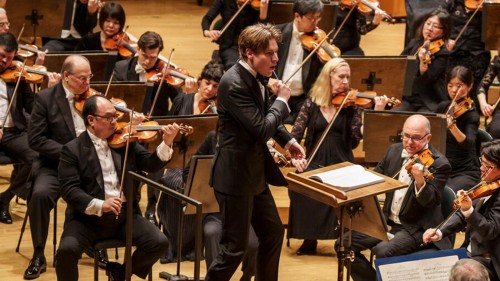 A young man conducts a large orchestra on a red podium; behind him the violin section is visible in formal concert attire. 