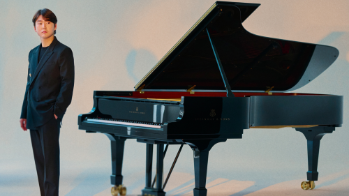 In a studio lit to resemble rosy sunlit clouds, a South Korean man in his 20s casually stands a few feet away from an open grand piano. he wears a dark suit and has his hands in his pockets.