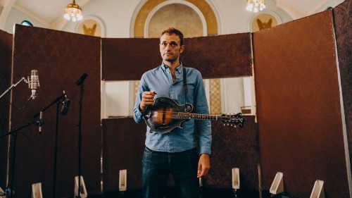 a mandolin player, in a recording booth within a historic church, pauses with his pick in hand and his instrument across his body