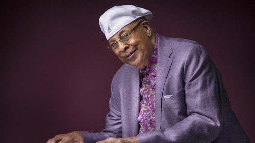 an older cuban man in a purple jacket and a flat cap smiles, with his hands poised over a keyboard
