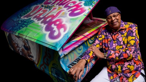 An older black man in a colorful purple, pink, and yellow top and a purple cap leans on a grand piano covered in colorful graffiti. 
