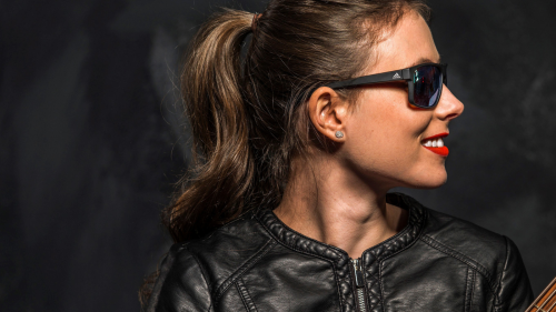 a young woman with dark glasses smiles as she turns her head to the neck of her bass guitar