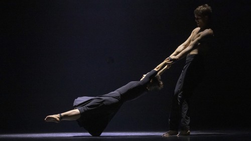 Two Circa performers wearing black against a black background, one stands and swings the other by their hands. 