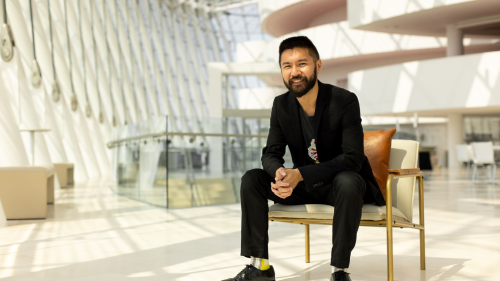 Conrad Tao, an Asian American man in his 30s, wears a slim black suit as he sits on a modern leather chair in a sun-filled atrium
