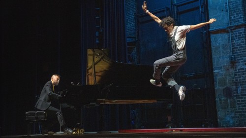 Two performers are pictured on a stage; one sits at a grand piano while the other is pictured high in the air, arms outstretched and tap shoes poised to land on a tapping surface. 