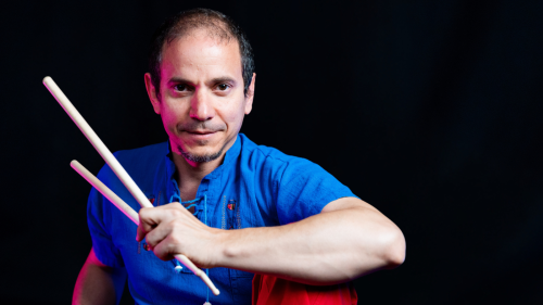 A Cuban American man holds a pair of drum sticks as he leans forward in front of a black background