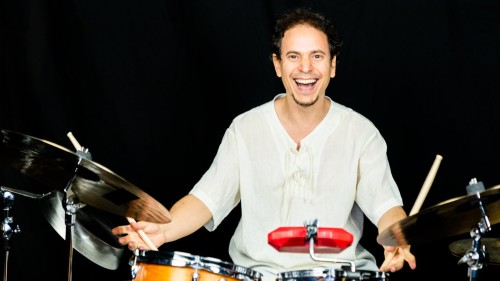 A Cuban American man sits behind a bright golden drum set, beaming in a white shirt in front of a black background. 