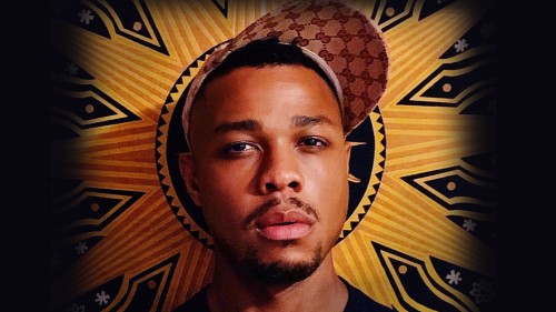 Davone Tines, a black man looks at the camera standing in front of an orange and black banner. 