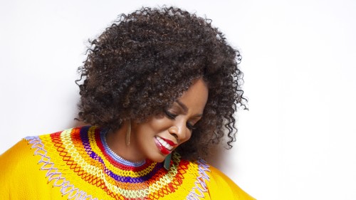 A black woman with shoulder length hair wears gold earrings and a bright yellow shirt with a colorful collar while looking downward. 