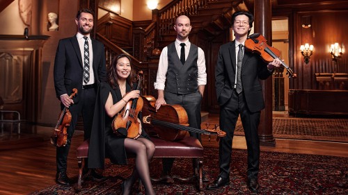 In a grand, wood paneled lobby with a staircase, a string quartet poses together in dark formal wear. A woman sits on a bench next to a horizontal cello holding a viola, while the three men stand behind her; the two outer men holding violins. 