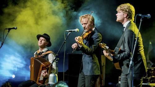 Three men perform on stage with string instruments and microphones. Yellow smoke billows behind them. 