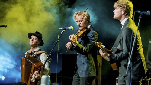 Three musicians stand diagonally, playing a guitar, violin, and accordion respectively in front of a greenish-blue fog. 