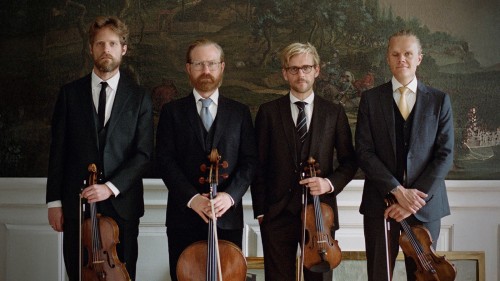 Four white men in a string quartet stand in a row in dark suits holding their instruments in front of several nature murals. 