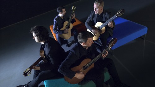 Four Irish guitar players sit spread out between three ottomans, facing different directions while wearing concert black. 