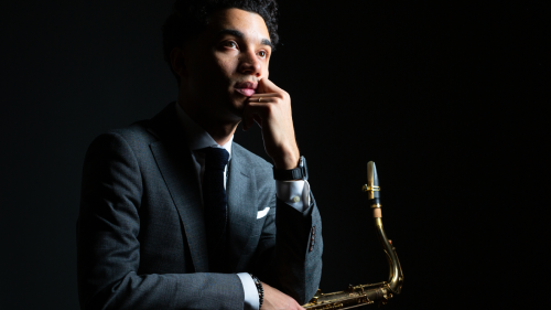A Puerto Rican man with dark, curly hair dressed in a gray suit sits against a black background, holding a tenor saxophone while leaning his chin on his left arm. 