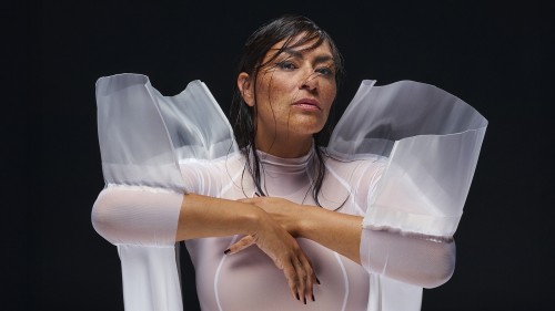 An Indigenous woman stands with her hands crossed in front of her in an ethereal white outfit, her hair strung across her face. 