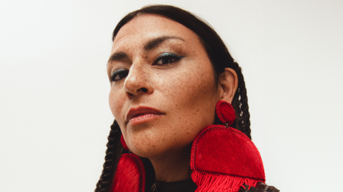 An Indigenous woman, in a 3/4 profile, looks down toward the camera. She wears red semicircular earrings made of fur with a beaded fringe.