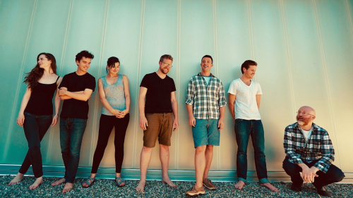 six musicians in casual summer clothes, four men and two women, stand in front of a ridged wall. another man crouches to their left.
