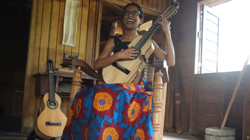 A young woman with short dark curls, glasses, and a bright floral skirt sits in a rocking chair smiling widely while holding a ten-string Puerto Rican cuatro. 