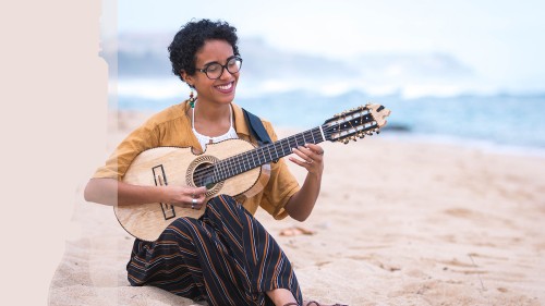 a young smiling puerto rican woman with curly short hair sits on the beach and plays the cuatro