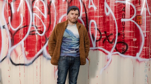 A white man stands in blue jeans, a t-shirt with mountains, and a brown fuzzy jacket in front of a wall of graffiti. 