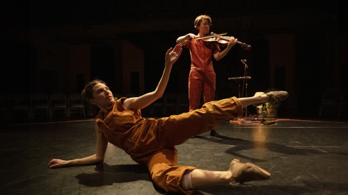 Two female performers appear in a black box space in burnt orange jumpsuits. One stands in the background playing the violin, while the other reclines with right elbow and knee on the ground, in an open outstretched position. 