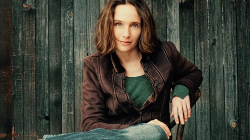 A green-eyed, brown-haired white woman. She smiles slightly and wears jeans, a brown suede jacket, and a green-blue wool sweater.