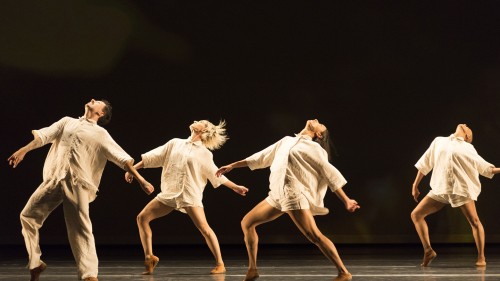 A dance company, men and women in ivory linen rompers, stepping forward, gesturing backward, and looking up in unison