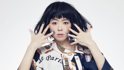 Hiromi, an Asian woman with dark hair wearing a patterned shirt holding her hands in front of her face in front of a white background. 