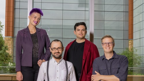 Four people stand in front of a shaded window. Two are standing and two are sitting.