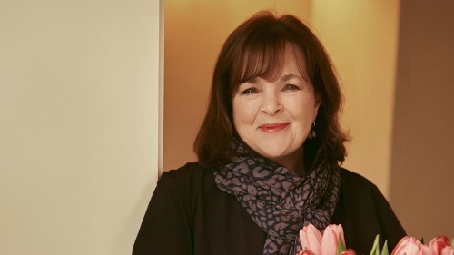 A portrait of an older, white woman with medium brown hair and bangs in a dark jacket with a dark pattered scarf, holding light pink tulips.  
