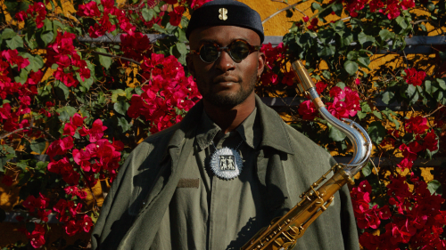 Isaiah Collier, a black man in his 20s, wears an olive green jumpsuit with an olive overcoat draped like a cloak, sunglasses, and a short lustrous cap