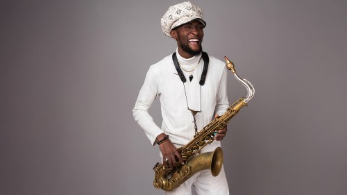 A young black man wearing an oversized paperboy cap stands holding a tenor saxophone with a vintage finish. 