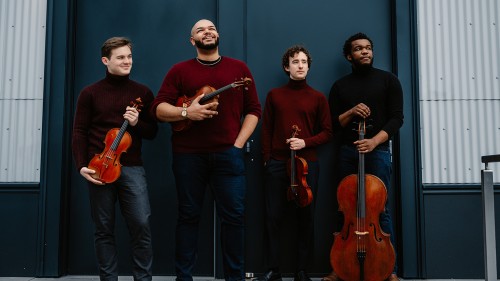 four young men smile impishly as they hold their string instruments in front of a bookstore with a bright blue facade.