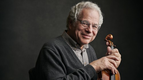 Violinst Itzhak Perlman, a man with grey hair wearing a grey sweater and button down against a gray background holds his violin. 