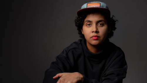 a dominican woman in her early 30s wearing a black sweatshirt and a baseball cap folds her arms across her chest as she kneels behind a drum