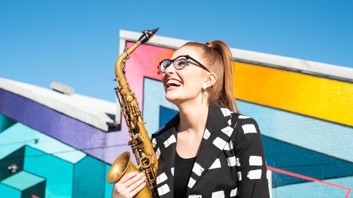 A young white woman with glasses and her red hair in a sleek ponytail, smiles broadly as she holds her tenor saxophone in front of a colorful geometric wall.