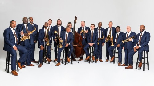 Members of Jazz at Lincoln Center Orchestra in blue suits with brown ties standing and sitting with their instruments against a white background. 