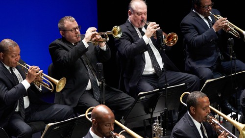 Four members of a jazz band are shown playing brass instruments in suits with polka dot ties on a stage with a blue background. 