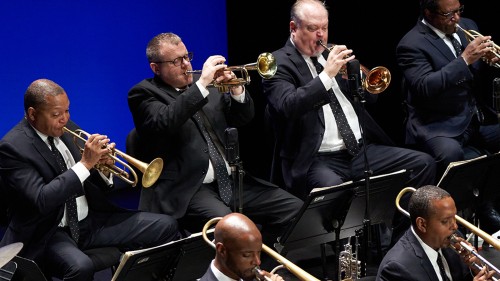 Four members of a jazz band are shown playing brass instruments in suits with polka dot ties on a stage with a blue background. 