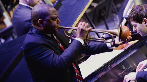 A grand piano with a white man playing fills the background of the image on an upward tilt, while a Black man plays trumpet with a mute in the foreground.  