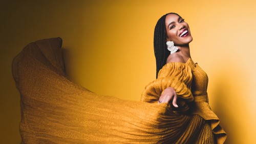 A black woman flourishes a pleated yellow dress while smiling widely in front of a shadowy yellow background. 